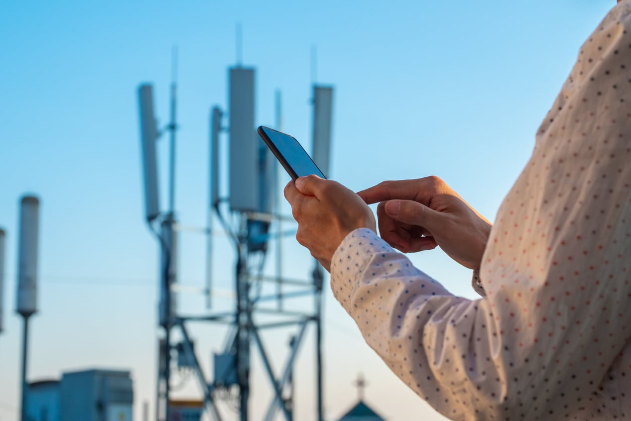 A person using a smartphone outside with a 5G tower in the background as emerging technologies..